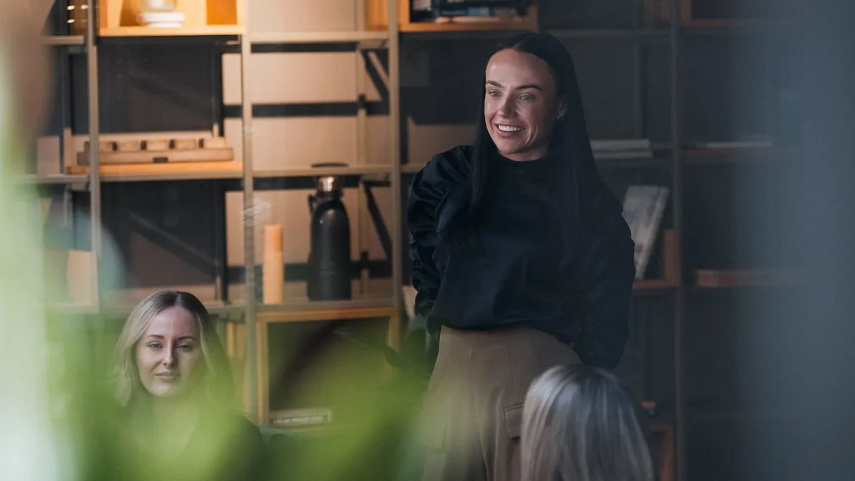 A woman stands and speaks in a cozy indoor setting with people seated nearby